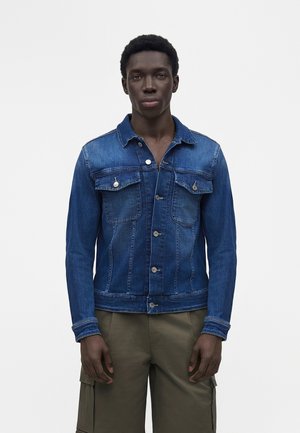 Mężczyzna stojący przodem, ubrany w niebieską dżinsową kurtkę i oliwkowe spodnie cargo na jednolitym białym tle.