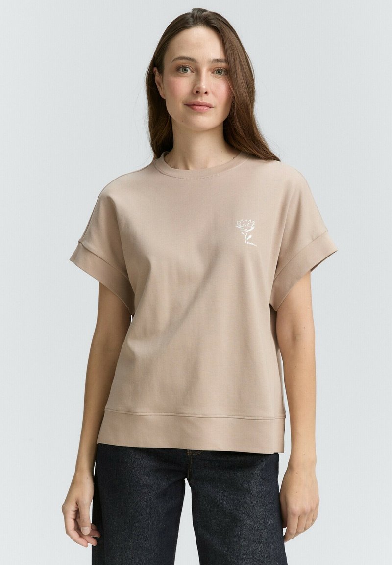 Sweatshirt beige clair avec des manches courtes retroussées, un col rond et un logo floral brodé sur la poitrine. Texture lisse, coupe décontractée.
