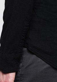 Primo piano del braccio di una persona che indossa una camicia nera a maniche lunghe con polsini abbottonati e pantaloni grigio scuro.