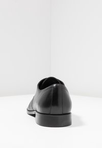 Chaussure oxford en cuir noir vue de l'arrière, montrant une finition lisse, des détails de couture et un petit talon sur une surface blanche.