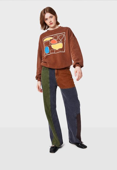 Brun oversized sweatshirt med abstrakt flerfärgad design, kombinerad med patchworkjeans i grönt, mörkblått och brunt. Svarta skor.