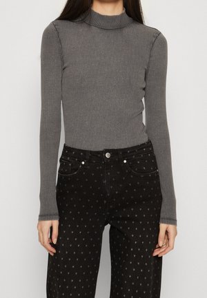 Frau trägt einen grauen, gerippten Langarm-Top mit Rollkragen, der in schwarze, hoch taillierte Hosen mit kleinen Sternmustern gesteckt ist.