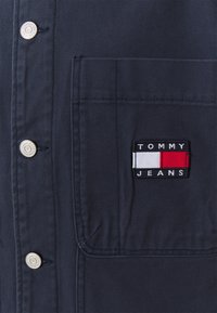 Marinblå skjorta med vävd textur, som har silverknappar och en vänster bröstficka med en rektangulär "TOMMY JEANS"-logotyp i röd, vit och svart.