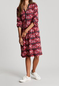 Czerwona sukienka w kwiaty z krótkimi rękawami, zapinana na guziki z przodu, z marszczoną talią i białymi trampkami. Charakteryzuje się żywymi kwiatowymi wzorami w odcieniach różu i zieleni.