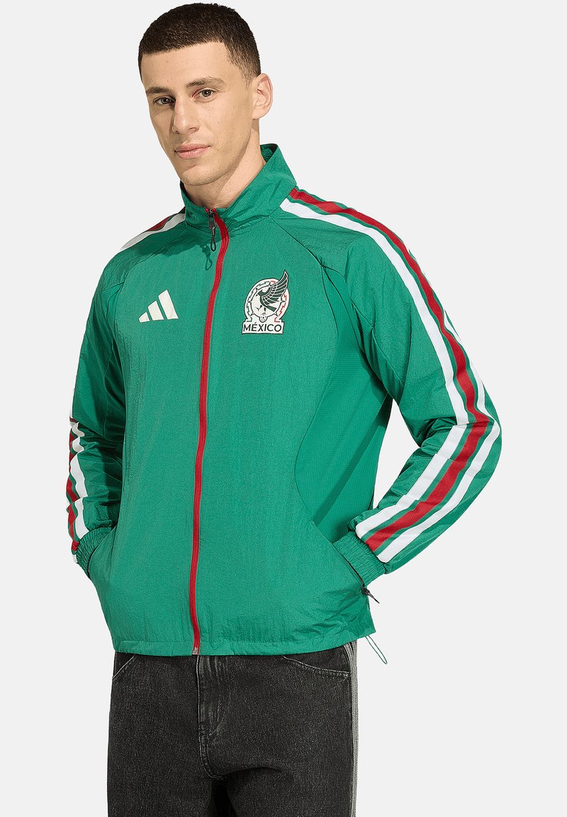 Man wearing a green Mexico sports jacket with red and white stripes on sleeves, Adidas logo, and hands in pockets against a plain background.
