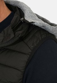 Gilet imbottito nero con cappuccio grigio, caratterizzato da una chiusura con zip e una superficie testurizzata. Il primo piano mostra l'area delle spalle e i dettagli del tessuto.