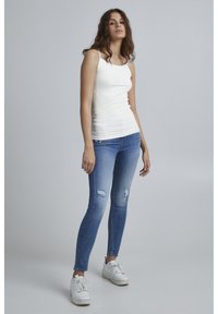 Weißes, enganliegendes Tanktop kombiniert mit hellblauen Skinny-Jeans mit ausgefranzten Akzenten, getragen mit weißen Sneakers. Einfaches und lässiges Design.