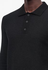 Polo noir texturé à manches longues avec col en tricot et trois boutons noirs sur la patte porté par une personne.