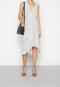 Robe blanche à pois noirs, décolleté en V, sans manches, ourlet asymétrique, associée à un petit sac noir et des sandales noires.