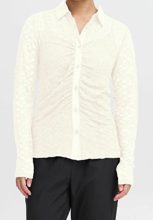 Wit gestructureerd overhemd met lange mouwen en kraag, gedragen met een zwarte broek. Handen ontspannen langs het lichaam.