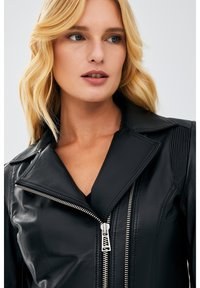 Zwarte leren jas met een ritssluiting aan de voorkant, brede kraag en geribbelde schouderdetails. Voorzien van zilveren ritshardware en een slank design.