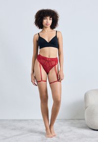 Bralette noire, ceinture porte-jarretelles en dentelle rouge avec des sangles. La ceinture présente un motif géométrique et des clips réglables. La mannequin se tient sur un tapis clair.