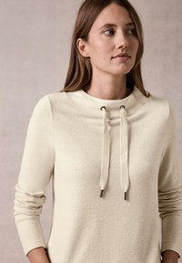 Beige Sweatshirt mit hohem Kragen, langen Ärmeln und Kordeln mit Metallenden. Die strukturierte Stoffqualität sorgt für ein weiches Aussehen.