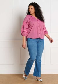 Mujer con cabello rizado que lleva una blusa rosa bordada, jeans azules y zapatos plateados puntiagudos, de pie frente a una pared blanca con paneles.