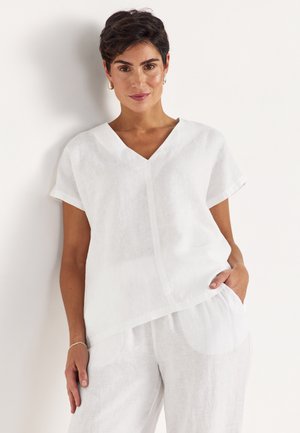 Kvinde med kort mørkt hår iført løs hvid V-hals hørtop og matchende hvide bukser, stående med den ene hånd i lommen.