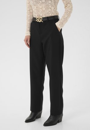 Schwarze maßgeschneiderte Hosen mit Seitentaschen, ein strukturierter Spitzenoberteil und ein goldfarbener Gürtel mit einer markanten S-förmigen Schnalle. Schwarze Stiefeletten mit Absatz.