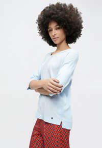 Jasnoniebieska bluzka z długim rękawem, zaokrąglonym dekoltem i bocznymi rozcięciami. Miękki materiał o subtelnej fakturze. Połączona z czerwonymi, wzorzystymi spodniami.