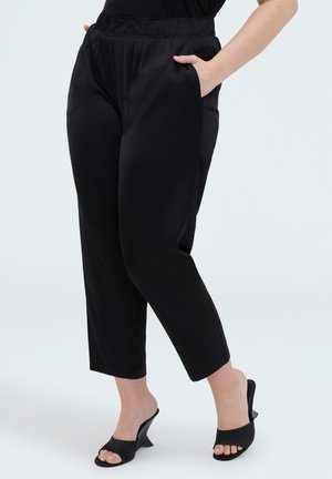 Frau trägt schwarze, gekürzte Hose mit elastischem Bund und schwarze, offene High-Heels mit Absatz, eine Hand in der Tasche.