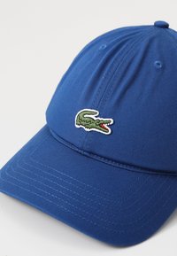 Casquette de baseball bleue en tissu texturé avec une visière incurvée. Présente un logo de crocodile vert brodé à l'avant.