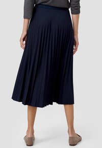 Femme portant une jupe midi plissée bleu marine, un haut à manches longues à motifs et des chaussures plates beiges, debout sur un fond uni.