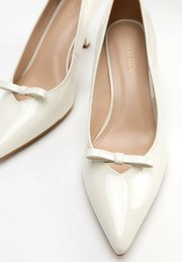 Scarpe con tacco alto in pelle verniciata bianca, con punta appuntita e dettaglio a fiocco decorativo sul davanti; tacco piccolo e fodera interna liscia.