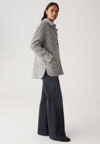 Frau, die eine grau-weiße strukturierte Jacke mit Taschen trägt, kombiniert mit hochtaillierten schwarzen weiten Jeans und schwarzen Schuhen, steht seitlich.