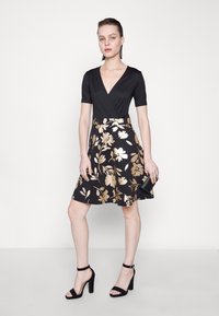 Robe noire à manches courtes avec un décolleté en V profond et une jupe évasée ornée de motifs floraux dorés. Associée à des talons noirs.