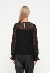 Mustapitsin blouse pitkillä hihoilla, jossa on avainaukko kaula-aukko ja röyhelömanchetit. Monimutkainen pitsikuvio, jossa on sileä, läpinäkyvä tekstuuri.