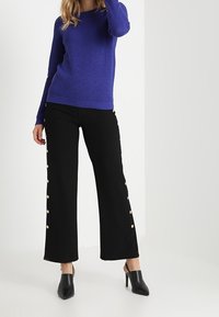 Blå texturerad tröja med långa ärmar, parat med svarta vida byxor med knäppdetaljer längs sidorna och svarta klackskor.