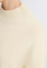 Pulover de culoare crem, cu guler înalt și nervuri, textură moale și croială lejeră, având umeri căzuți și detalii de design minimalist.