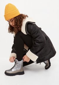 Bottines argentées métalliques avec fermeture éclair à l'avant, semelle épaisse noire. Manteau noir avec un col en polaire crème, leggings noirs et un bonnet en tricot jaune.