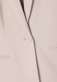 Blazer beige clair avec une texture lisse, fermeture à un bouton, revers crantés et deux poches avant avec des accents de couture.