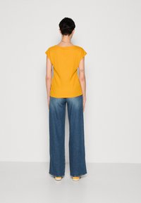 Person med kort hår som bär en gul kortärmad topp, blå vida jeans och gula skor, stående vänd mot en enfärgad vit vägg.