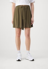 Olivově zelené se slim fit šortky s texturovanou látkou, elastickým pasem a detailem na zavazování vpředu. Noseny s bílými sportovními teniskami.