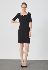 Robe noire ajustée avec des manches courtes, décolleté carré et coutures détaillées. Portée avec des escarpins noirs et un bracelet métallique. Design simple.