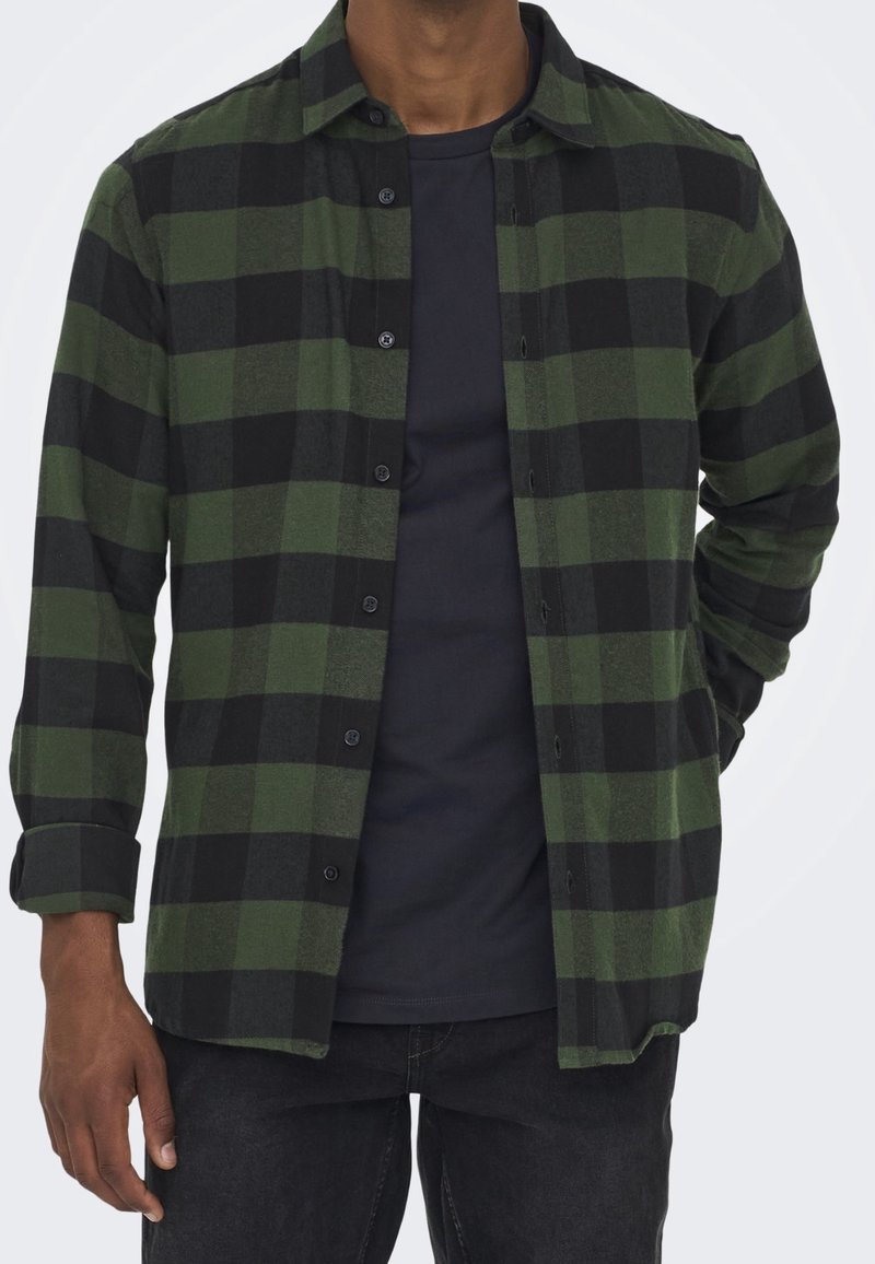 Chemise à carreaux verte et noire en flanelle, dotée de boutons, d'un col et de manches longues, portée par-dessus une chemise foncée.