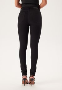 Czarne wąskie jeansy o gładkiej fakturze, z wysokim stanem i tylnymi kieszeniami, w połączeniu z czarnymi sandałami na wysokim obcasie. Widok z tyłu.