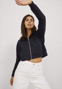 Hoodie cropped azul-marinho com fecho à frente, duas bolsos laterais e capuz com cordão, combinado com jeans brancos de cintura alta. Textura suave.