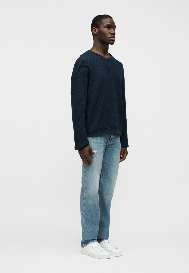 T-shirt à manches longues en maille côtelée bleu marine, coupe décontractée, associé à un jean bleu clair à jambes droites avec des détails subtilement usés et des baskets blanches.