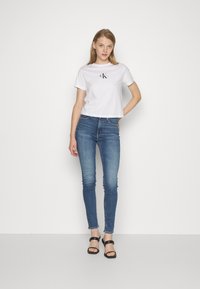 Bílý zkrácený tričko s černým logem "CK", spárované s modrými slim džíny. Modelka má na sobě černé sandály na pásku a stojí na jednobarevném pozadí.
