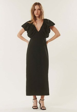 Femme portant une longue robe noire avec des manches longues à volants et bouffantes, un décolleté en V profond, des talons à bride à la cheville et un court collier de perles, debout avec les mains sur les hanches.