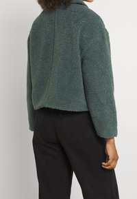 Grøn fleecejakke med en struktureret overflade, kort design og brede ærmer, kombineret med sorte denimbukser. Bagsiden viser sømdetaljer.