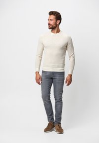 Hellbeige Pullover mit langen Ärmeln, figurbetontem Design, kombiniert mit grauen Jeans und braunen Sneakers, vor einem einfarbigen Hintergrund stehend.