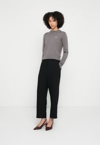 Maglione grigio lavorato a maglia con scollo a costine, pantaloni a gamba larga neri e décolleté bordeaux con punta affilata. La modella posa davanti a uno sfondo bianco semplice.