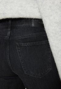 Lähikuva mustast denimist valmistatud teksapükste tagataskust ja vööst, koos heledat hallist kudumist valmistatud kampsuniga.