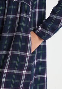 Vestido de cuadros azul marino hecho de franela, con patrones de cuadros en púrpura y verde, mangas largas y bolsillos laterales para mayor utilidad.