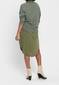 Nainen, jolla on päällään vihreä neule, oliivinvihreä hame, jossa on sivureiät, ja beige korkokenkä, seisoo yksinkertaista taustaa vasten.