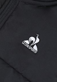 Tissu matelassé noir avec un logo brodé d'un coq stylisé et le texte "LE COQ SPORTIF" en dessous, en blanc.