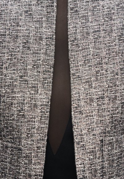 Detailní záběr na přední otvor texturovaného černobílého kostkovaného saka přes průhlednou černou látku a černý oděv pod ní.