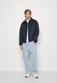 Giubbotto imbottito blu navy con texture trapuntata, dotato di chiusura a scatto, indossato sopra una maglietta bianca e abbinato a jeans azzurri chiari e sneakers.